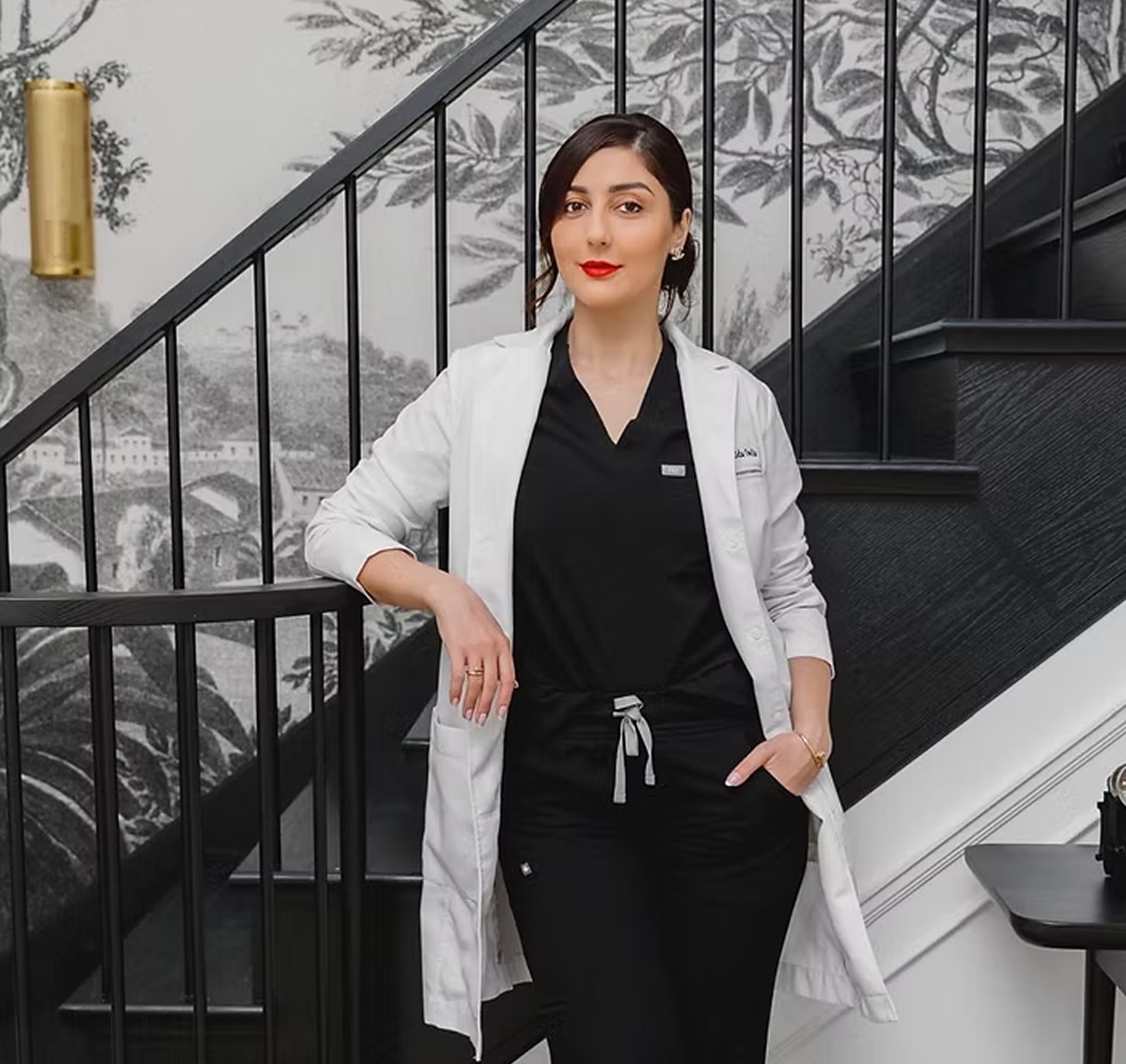Contact Dr. Owlia Aesthetics — Dr. Aida Owlia in a white coat and black scrubs beside a staircase